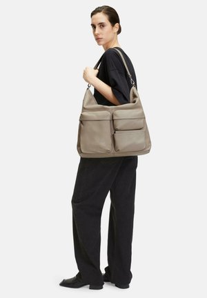 Beige leather shoulder bag with two front zippered pockets and a soft, structured design. The model is wearing a loose black top and wide-leg pants.