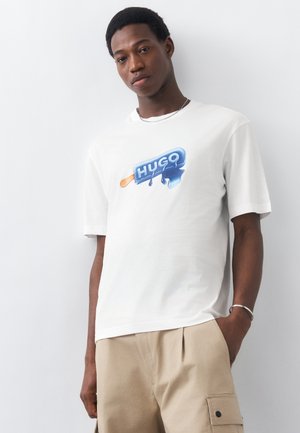 Άνδρας που φοράει λευκό T-shirt με μπλε σχέδιο λιωμένου παγωτού και κείμενο "HUGO", συνδυασμένο με μπεζ παντελόνι cargo, στέκεται μπροστά σε λευκό τοίχο.