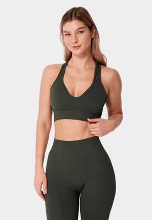 Kobieta o długich jasnobrązowych włosach, ubrana w ciemnozielony sportowy stanik i legginsy z wysokim stanem, pozująca z prawą ręką przy talii.