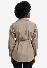 Beige cotton shirt with long sleeves, a back belt detail, and a collar. Features buttoned cuffs and pleats at the back for added shape.