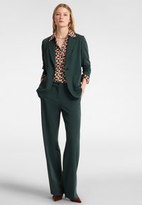 Vrouw staat met haar handen in de zakken, gekleed in een donkergroen pak met brede pijpen en een gebloemde blouse met geometrische vormen.