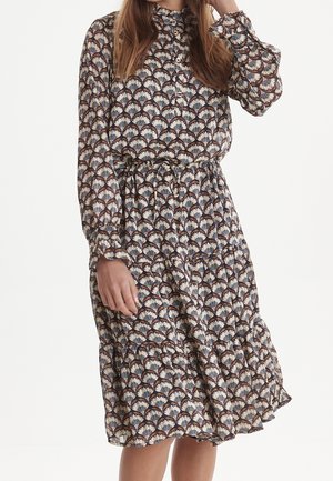 Femme portant une robe midi à manches longues boutonnée avec un motif floral en forme d’éventail marron, crème et marine et une taille avec cordon.