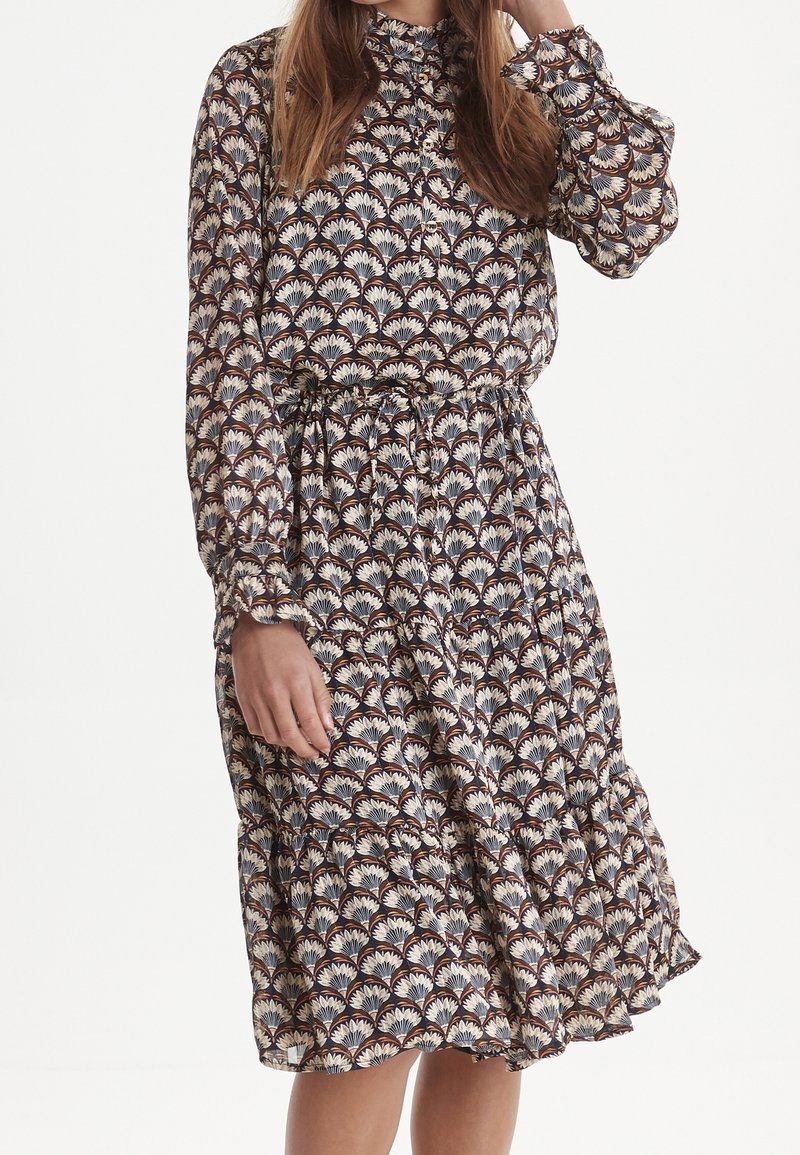 Femme portant une robe midi à manches longues boutonnée avec un motif floral en forme d’éventail marron, crème et marine et une taille avec cordon.