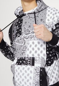 Mikina s kapucí s patchworkovým designem, který obsahuje černé, bílé a šedé paisley vzory. K dokončení vzhledu přispívají stahovací šňůrky a žebrované manžety.