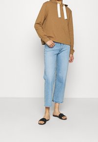 Felpa marrone con cappuccio e cordini bianchi, jeans blu a gamba dritta e sandali neri a ciabatta. Vestibilità casual e rilassata con tessuto morbido.