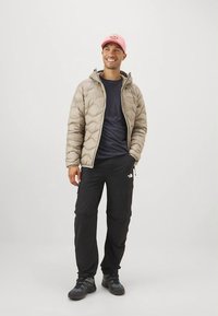 Lächelnder Mann, der eine beigefarbene Puffjacke, schwarze Hose, schwarze Schuhe und eine pinkfarbene Baseballmütze trägt, mit einer Hand in der Tasche stehend.
