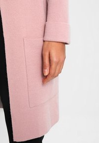 Vaaleanpunainen neulottu neuletakki, jossa on käännetty hihansuu ja edessä suorakulmainen tasku, sekä pehmeä ja sileä pinta.