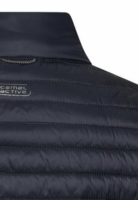 Nahaufnahme des Rückens einer marineblauen, gesteppte Jacke mit hohem Kragen und dem "Camel Active"-Logo auf der linken Seite.