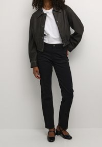 Giubbotto corto in pelle nera, t-shirt bianca, jeans neri a vita alta e scarpe con tacco nere. Il giubbotto presenta bottoni a pressione e un ampio colletto.