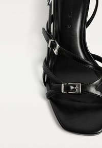 Zwarte hoge hak sandalen met bandjes en een vierkante neus. Voorzien van glanzende zilveren gespen en een gladde leren textuur. Minimalistisch ontwerp en slanke vorm.