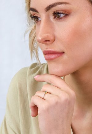 Gold-tone ring with a small green gemstone, worn on a hand resting near a face. Soft lighting highlights the ring's details.