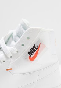 Valkoiset korkeavartiset tennarit, jotka on valmistettu teksturoidusta kankaasta. Niissä on erottuva punainen Nike-logotagi ja oranssi koristeompelu sivussa.