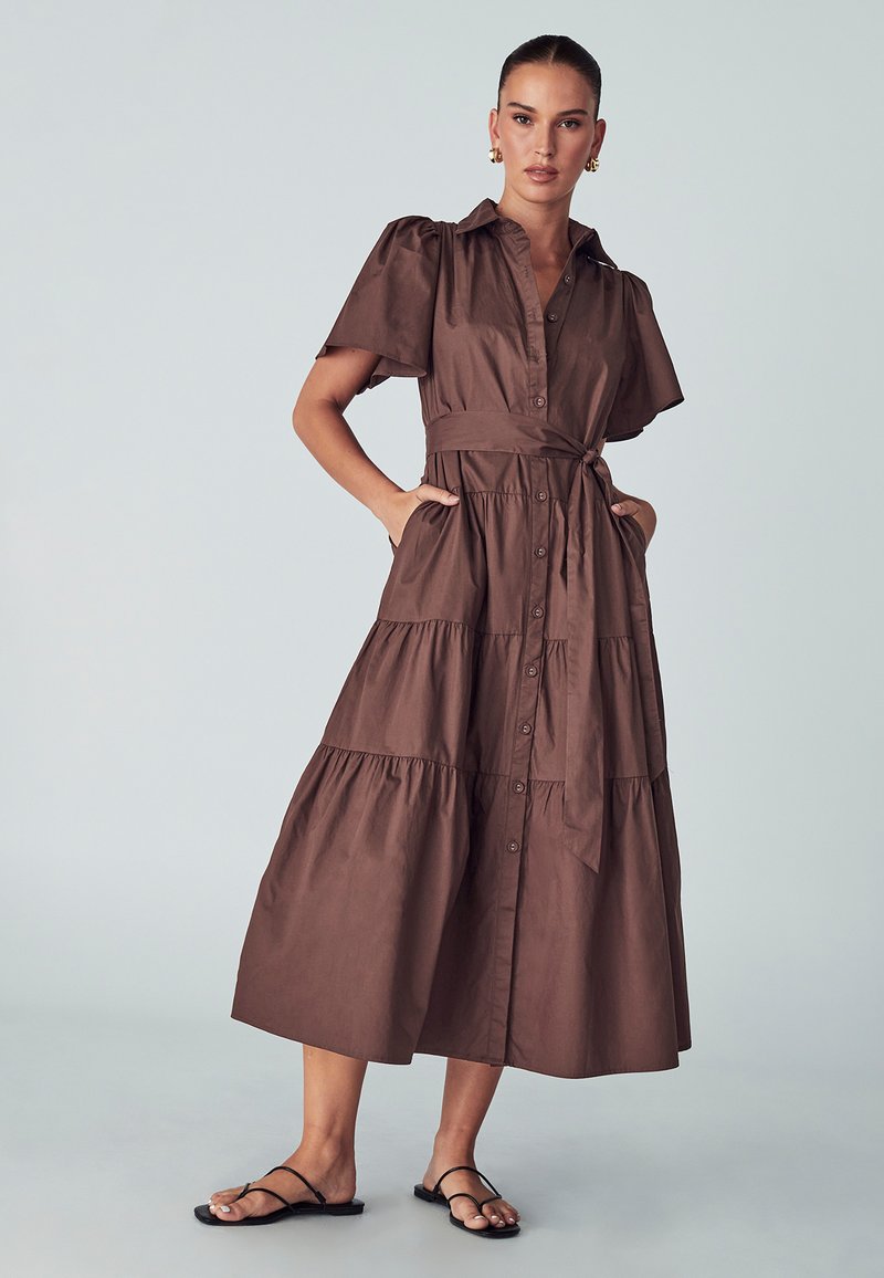 Rjava bombažna obleka s kratkimi napihnjeni rokavi, z gumbi spredaj in pasom. Ima zasnovo večplastne krila in stranske žepe. Obuti so ravni sandali.