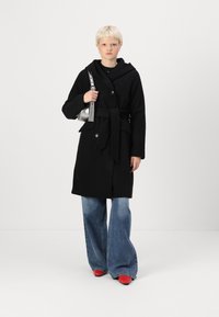 Abrigo de lana negro con capucha, cintura ceñida con cinturón, complementos en dorado, combinado con jeans acampanados de color azul claro y zapatos rojos de punta. Acentos en plateado en el bolso.