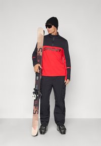 Rood en zwart fleece top met ritssluiting en logo, zwarte skibroek, zwarte beanie en zonnebril; houdt een paar ski's met bindingen vast.