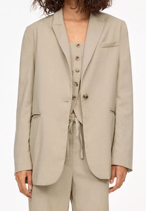 Woman wearing beige pinstripe blazer, matching buttoned vest, and drawstring pants, shown from mid-chest to mid-thigh.