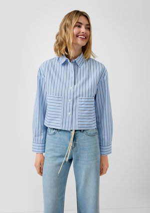 Femme souriante portant une chemise à rayures verticales bleues et blanches avec deux poches avant à rayures horizontales et un jean bleu clair avec un cordon de serrage.