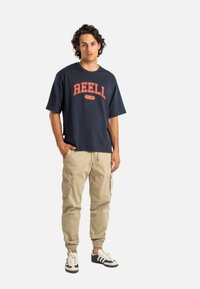 Joven con cabello rizado que lleva una camiseta REELL de color azul marino, pantalones cargo beige y zapatillas blancas con rayas azules, de pie con las manos en los bolsillos.