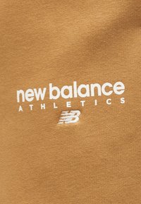 Μπεζ ύφασμα με λευκό κείμενο «new balance ATHLETICS» και το λογότυπο της New Balance κεντημένο κάτω από το κείμενο.