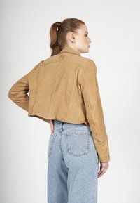 Tan suedine kort jasje met een platte kraag, zichtbare stikdetails en lange mouwen, gecombineerd met hoge taille lichte denim jeans.