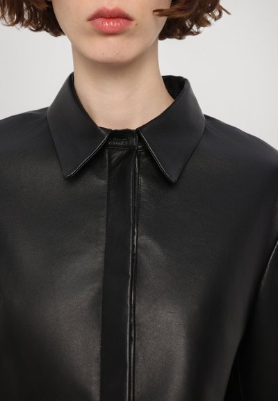 Črna usnjena srajčna jakna s klasičnim ovratnikom, ki se ponaša z gladko teksturo in sprednjim zadrgo, kar poudarja dovršen izgled.