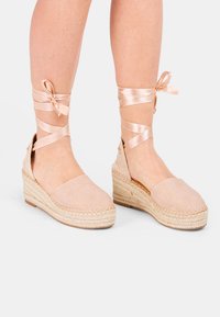 Bāli rozā espadrilles ar augstiem papēžiem, ar teksturētām jūtas zolēm, mīksto auduma augšdaļu un samta lentēm, kas sasienas ap potīti, nodrošinot regulējamu atbalstu.