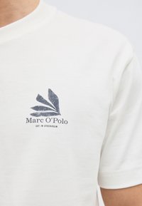 Λευκό μπλουζάκι με μικρό μαύρο λογότυπο Marc O’Polo και αφηρημένο σχέδιο φύλλου στο αριστερό στήθος, φορεμένο από άτομο περικομμένο από τον λαιμό έως τη μέση.