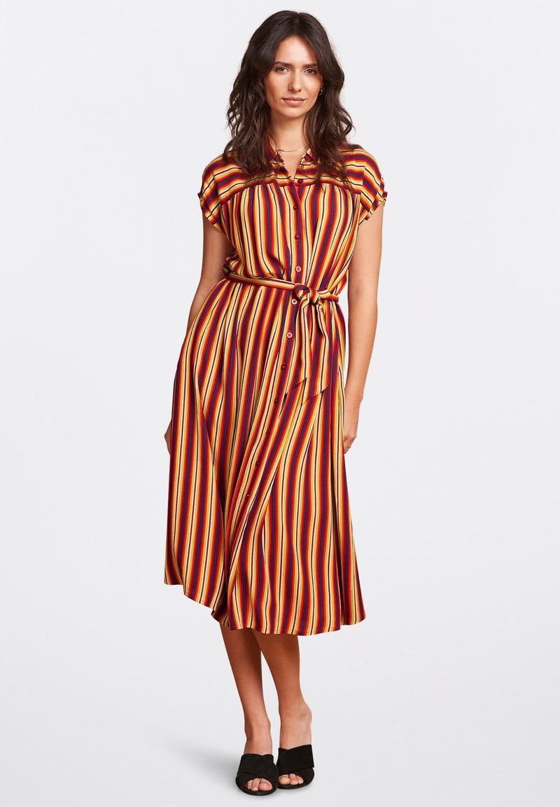 Vestito a camicia a righe in rosso, arancione e giallo; vita con cintura; maniche corte, orlo fino al ginocchio; abbinato a sandali neri con punta aperta.