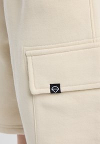 Beige shorts with a smooth texture, featuring a front patch pocket and a black label with a white logo on the side.