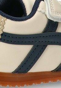 Zapato beige claro con rayas azul marino, parte superior de cuero texturizado, detalles de cordones beige y suela de goma marrón. Presenta un detalle de costura en contraste.