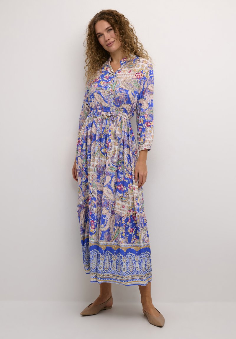 Dolga modra obleka z vzorcem paisley in cvetličnimi akcenti, ovratnikom in zategnjenim pasom. Tkanina se zdi lahka in zračna.