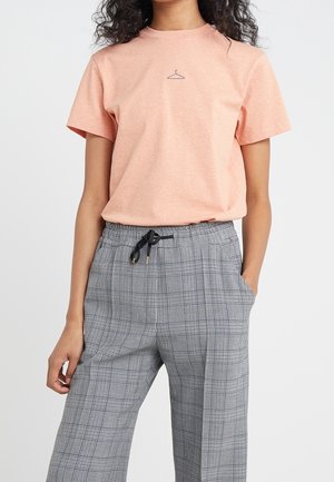 Woman wearing a peach t-shirt with a small hanger icon and high-waisted gray plaid pants with a black drawstring and pockets.