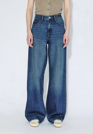 Person wearing high-waisted, wide-leg blue jeans with a button and zipper fly, paired with white sneakers and a beige sleeveless top.