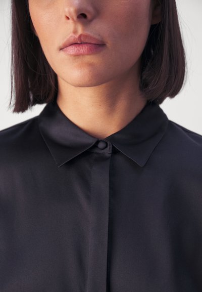 Chemise noire à boutons avec un col classique. Le tissu semble lisse et légèrement brillant, mettant en valeur un design épuré.