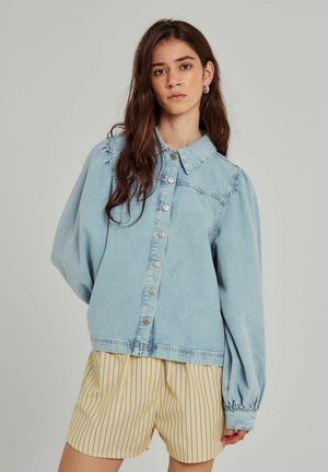 Lichtblauwe denimjas met een knoopsluiting aan de voorkant, puntige kraag en gathered mouwen. Gecombineerd met gele pinstripe shorts.