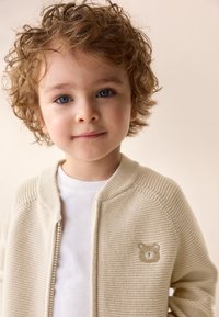 Beige knitted cardigan with a ribbed texture and embroidered bear face detail. Worn over a white t-shirt, featuring a zipper closure.