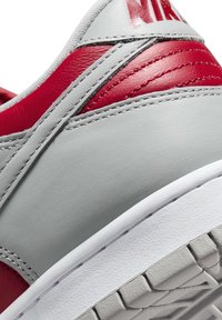 Nike-sneakerdetalj med en slät grå läderöverdel, röda accentpaneler och vit gummisula. Har strukturerade linjer och sömmar.