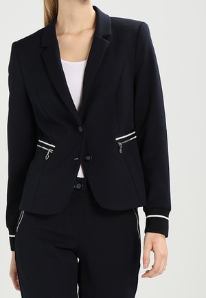 Kvinde iført en tætsiddende sort blazer med hvide lynlåslommer, hvide kanter på ærmerne og matchende sorte bukser med hvide sidelommer.