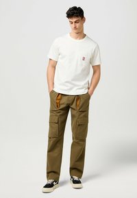 Hvid bomulds T-shirt med en brystlomme og logo. Olivengrønne cargo-bukser med orange accenter og flere lommer. Sorte sneakers med hvide såler.