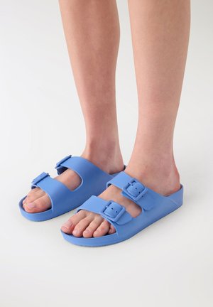 Piedi che indossano sandali ciabatta azzurri a doppia fascia con fibbie regolabili su sfondo bianco semplice.