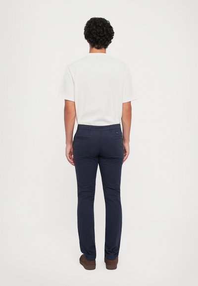 Uomo in piedi di spalle, che indossa una camicia bianca a maniche corte, pantaloni blu scuro e scarpe marroni su uno sfondo chiaro e semplice.