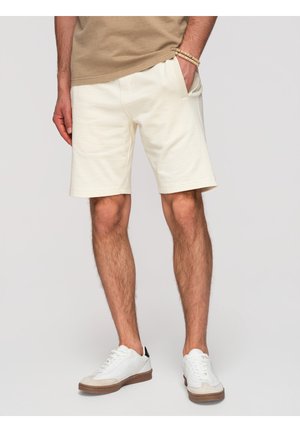 Mann trägt beige Shorts, weiße Sneaker und ein braunes T-Shirt mit einer Hand in der Tasche vor schlichtem Hintergrund.