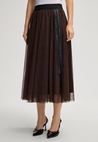 Mulher vestindo uma saia midi de tule castanha com um cinto preto da marca Joop! e sapatos de salto alto pretos com bico fino e tiras no tornozelo.