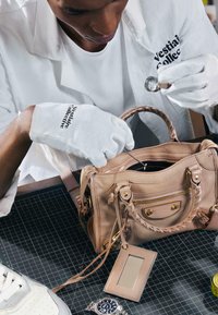 Personne portant des gants blancs examine un sac à main beige avec une loupe et une épingle sur un tapis de découpe noir.