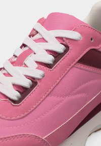 Κοντινή λήψη ενός ροζ αθλητικού παπουτσιού με λευκά κορδόνια, λεπτομερή ραφές και διχρωμία υφής στο άνω μέρος, σε λευκό φόντο.