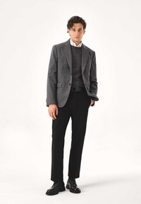Blazer grigio con revers a incavo, indossato sopra un maglione scuro. Pantaloni neri e scarpe nere lucide completano l'outfit.