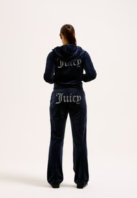 Jūras spēku zilo velūra sporta tērps ar kapuci, kas sastāv no jakas un biksēm. Vārds "Juicy" ir greznoti ar rhinestones gan uz jakas, gan uz biksēm.