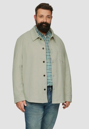 Lichtgrijze button-up jas met een kraag, gedragen over een blauw-groen geruit shirt. Bevat contrasterende donkere knopen en een borstzak.