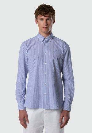 Uomo che indossa una camicia a righe verticali blu e bianche con bottoni e maniche lunghe, e pantaloni bianchi, in piedi contro uno sfondo semplice.