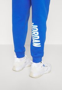 Modré tepláky s nápisem "JORDAN" tištěným bílou barvou vertikálně na nohavici. Spárováno s bílými sportovními botami s texturovaným designem.